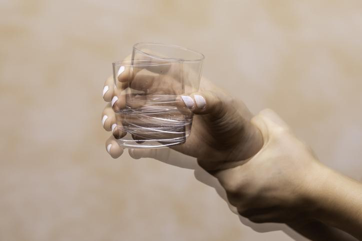 A shaky hand holding a glass & a second hand gripping wrist for support; two images of the glass show against peach background.