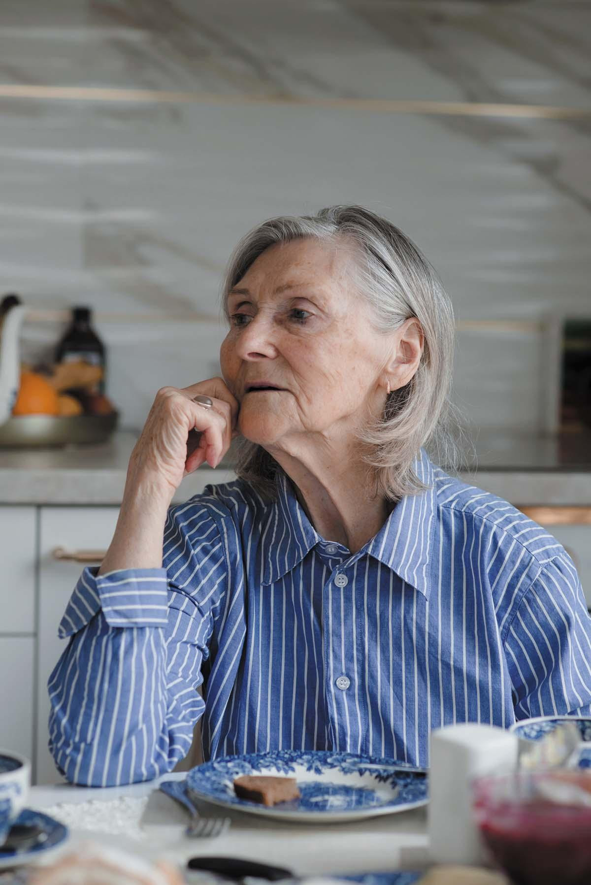 A mature woman sits at a table with a piece of toast on a plate in front of her, looking like she is deep in thought..