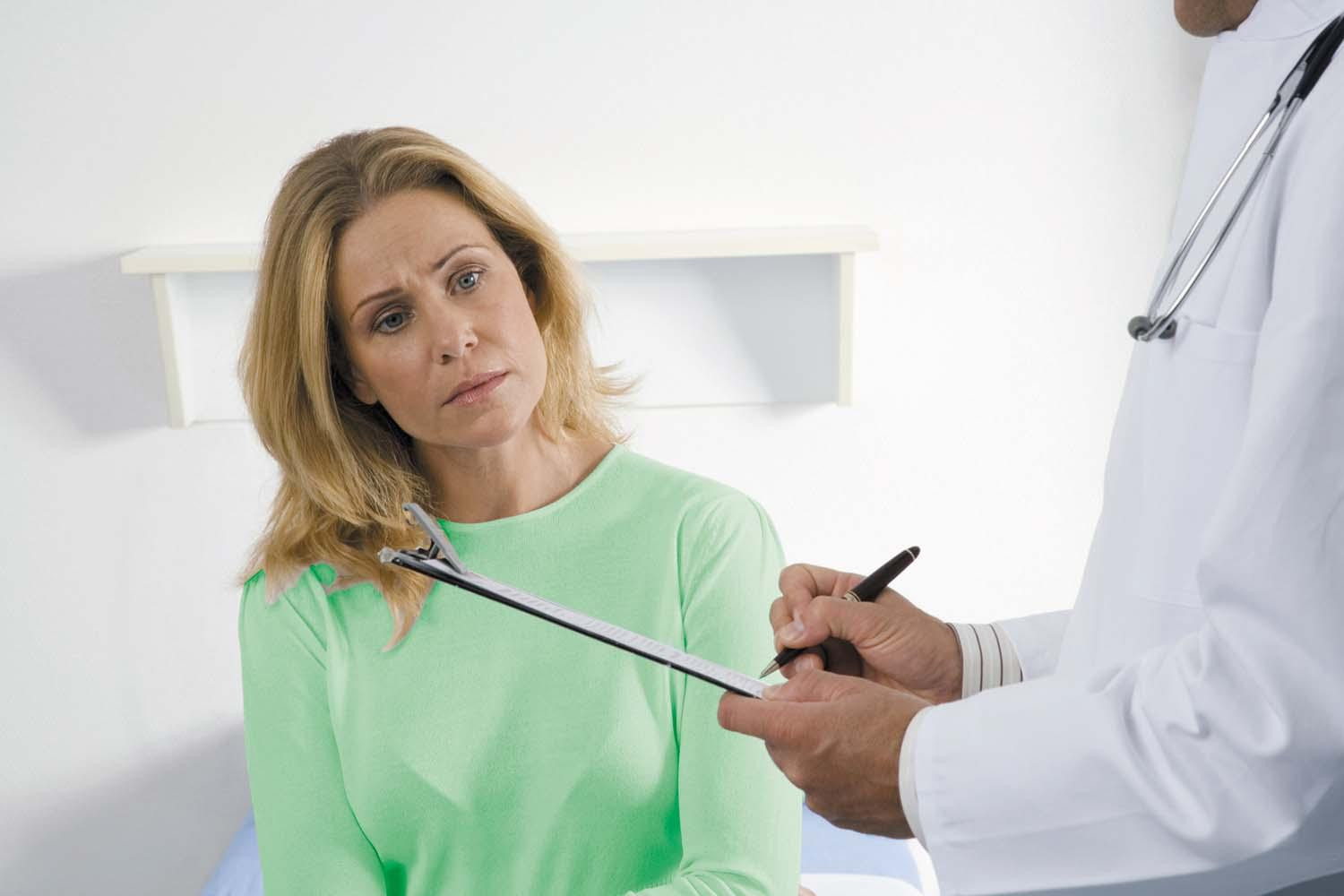 A woman in a doctor's office sitting next to the doctor as he writes on a clipboard.